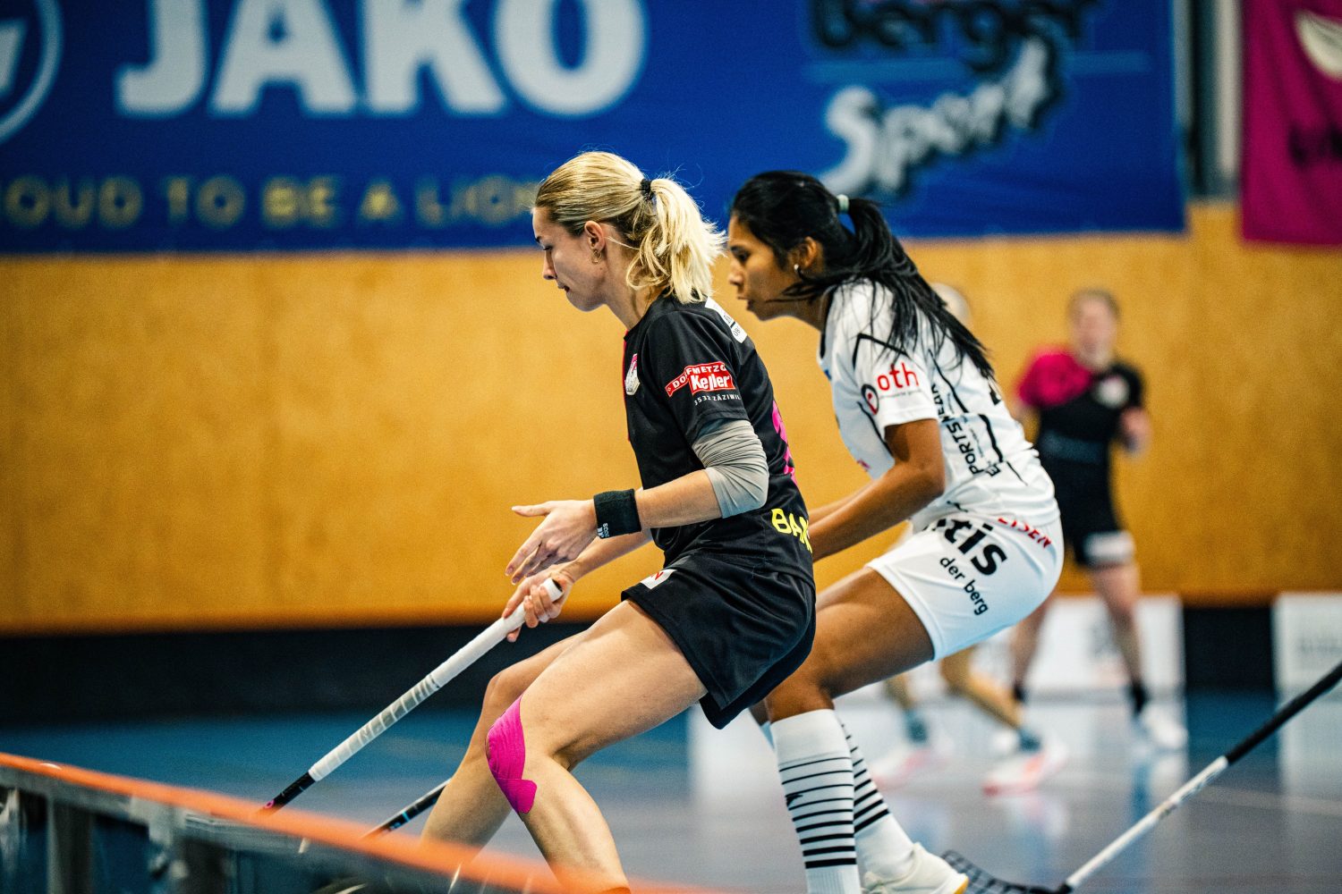 Zwei Frauen, Soraya und Leonie, treten bei einem Unihockeyspiel in einer Halle gegeneinander an, wobei beide darauf bedacht sind, den Ball mit ihren Schlägern zu kontrollieren. Eine Spielerin trägt schwarz, die andere weiß, und im verschwommenen Hintergrund ist ein Sporthallenbanner zu sehen.