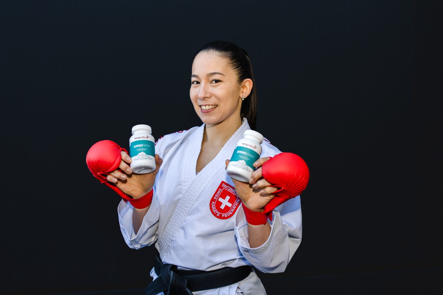 Ein lächelnder Kampfsportler in einem weißen Kampfanzug mit rotem Schweizer Abzeichen und roten Boxhandschuhen hält zwei Flaschen Amino Vida vor einem schlichten dunklen Hintergrund hoch.