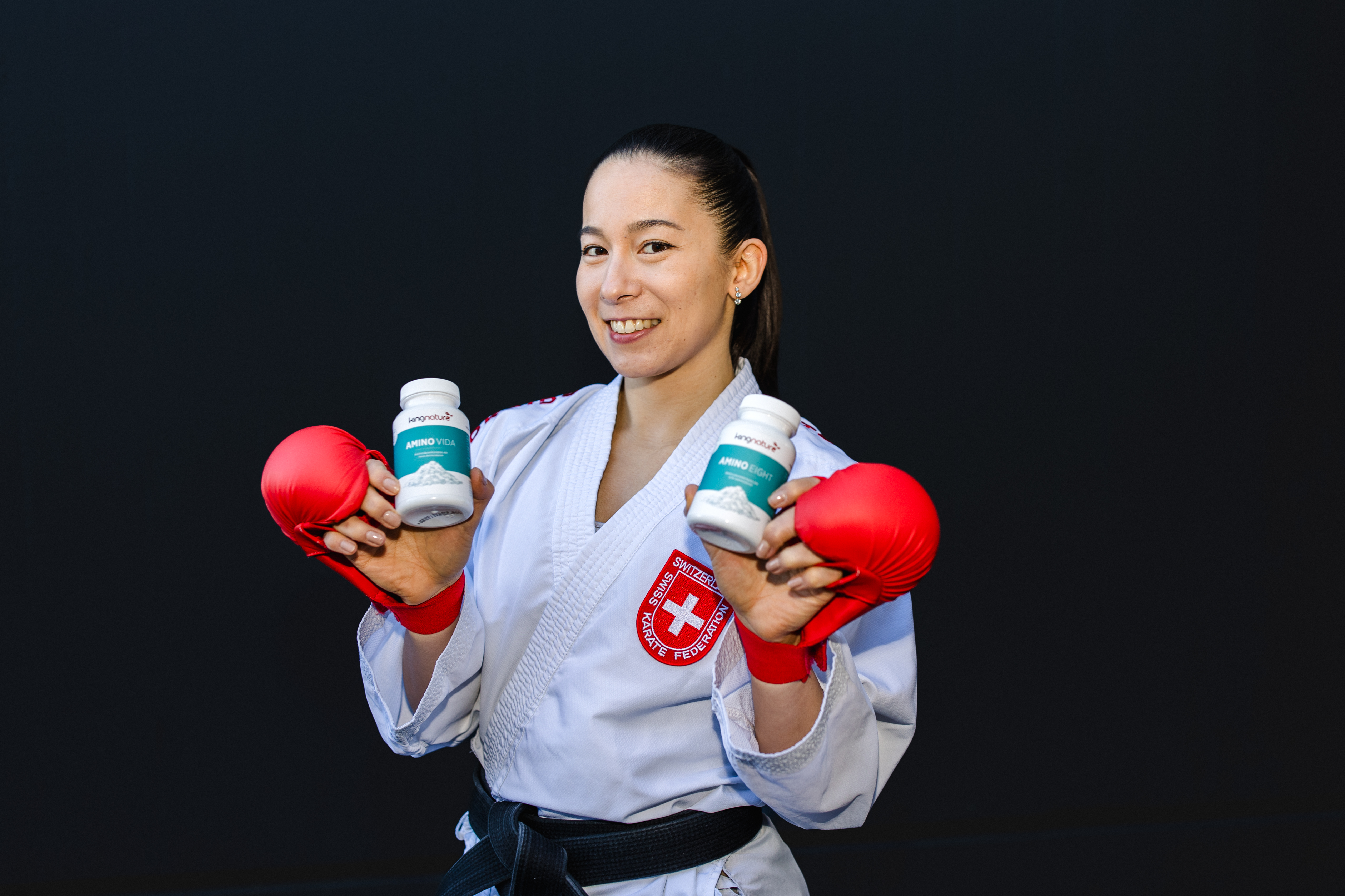 Ein lächelnder Kampfsportler in einem weißen Kampfanzug mit rotem Schweizer Abzeichen und roten Boxhandschuhen hält zwei Flaschen Amino Vida vor einem schlichten dunklen Hintergrund hoch.