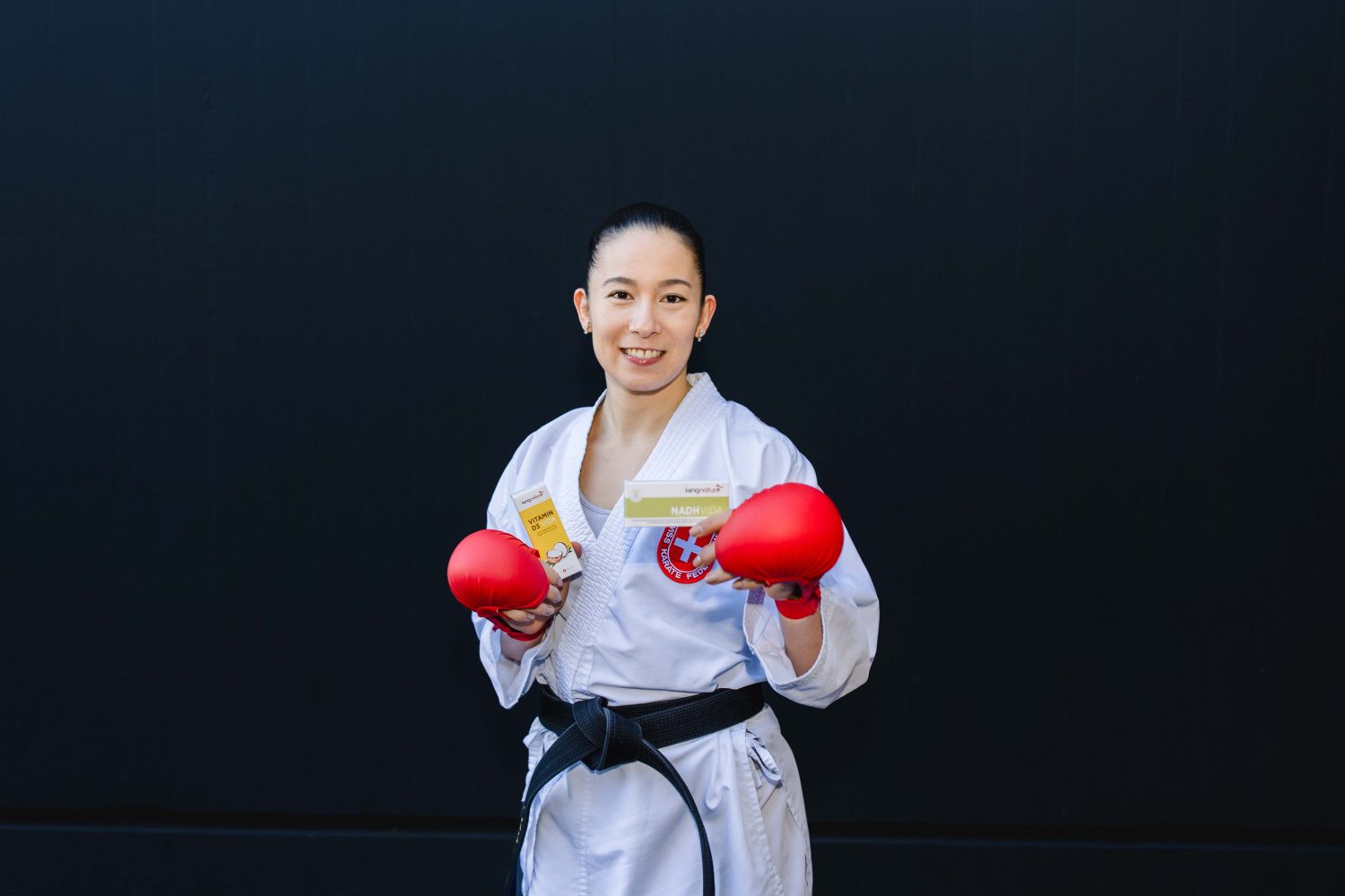 Eine Sportlerin in einem rot-weißen Sportoberteil und schwarzen Shorts kniet in einer Sprint-Startposition und lächelt vor einem schlichten schwarzen Hintergrund in die Kamera - und verdeutlicht so den Schub, den Sie mit Aronia Shot spüren.