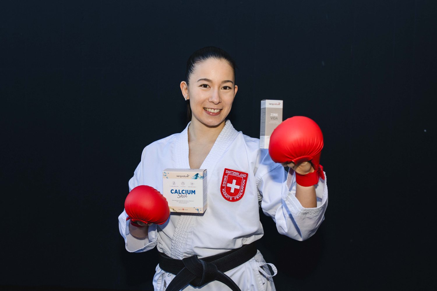Eine Frau in einem Karateanzug mit Schweizer Aufnäher lächelt, trägt rote Handschuhe und hält Zink Vida und eine Calcium-Dose in der Hand. Sie wirbt für Zink-Tropfen kaufen vor einem dunklen Hintergrund.