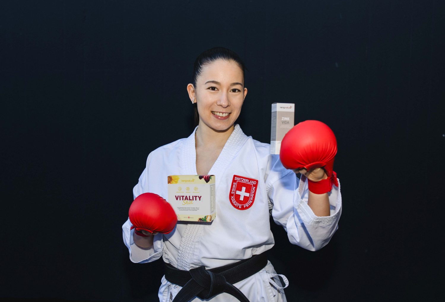 Eine Frau in Karateuniform und roten Handschuhen lächelt, während sie vor einem dunklen Hintergrund eine Schachtel mit der Aufschrift Vitality Shot in der Hand hält.