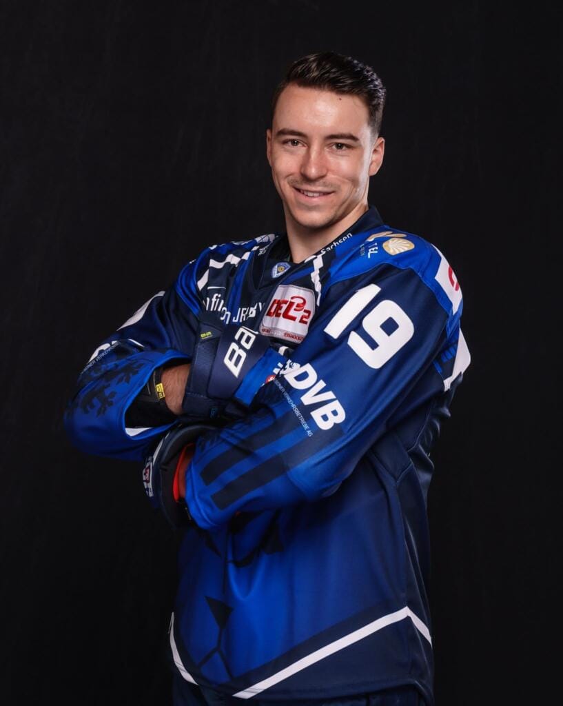 Niklas Postel, ein männlicher Hockeyspieler im blauen Trikot mit der Nummer 19, steht mit verschränkten Armen und lächelt selbstbewusst vor einem schwarzen Hintergrund.