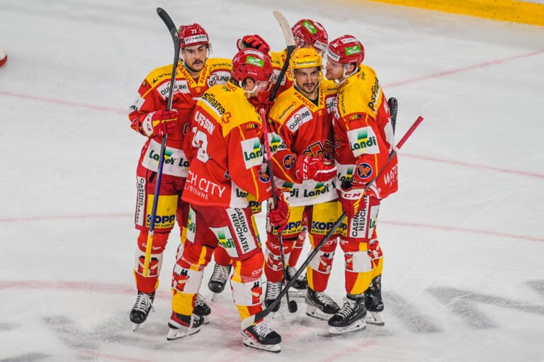 Fünf engagierte Eishockeyspieler in roten und gelben Uniformen drängen sich auf dem Eis und feiern. Sie halten Hockeyschläger, während im Hintergrund das Spitzen-Eishockey auf der Eisbahn mit gelber Begrenzung zu sehen ist.