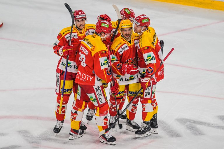 Fünf Eishockeyspieler des EHC Biel in rot-gelben Trikots versammeln sich auf dem Eis und feiern einen Spielzug. Sie stehen dicht beieinander in der Mitte der Eisfläche und zeigen grossen Teamgeist, während sie sich auf den Schlussspurt vorbereiten.