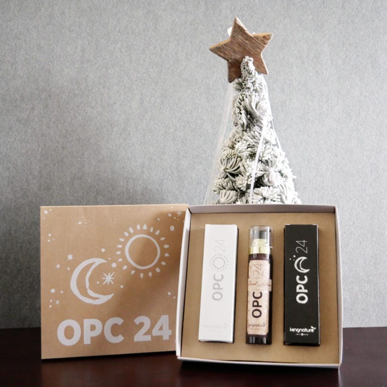 Eine weihnachtliche OPC 24 Geschenkbox mit einem Dreierset beschrifteter Artikel, darunter eine Tube und zwei rechteckige OPC-Creme-Boxen, ist geöffnet und steht vor einem kleinen weiß-goldenen Weihnachtsbaum mit Stern als Spitze. Der Hintergrund ist grau.