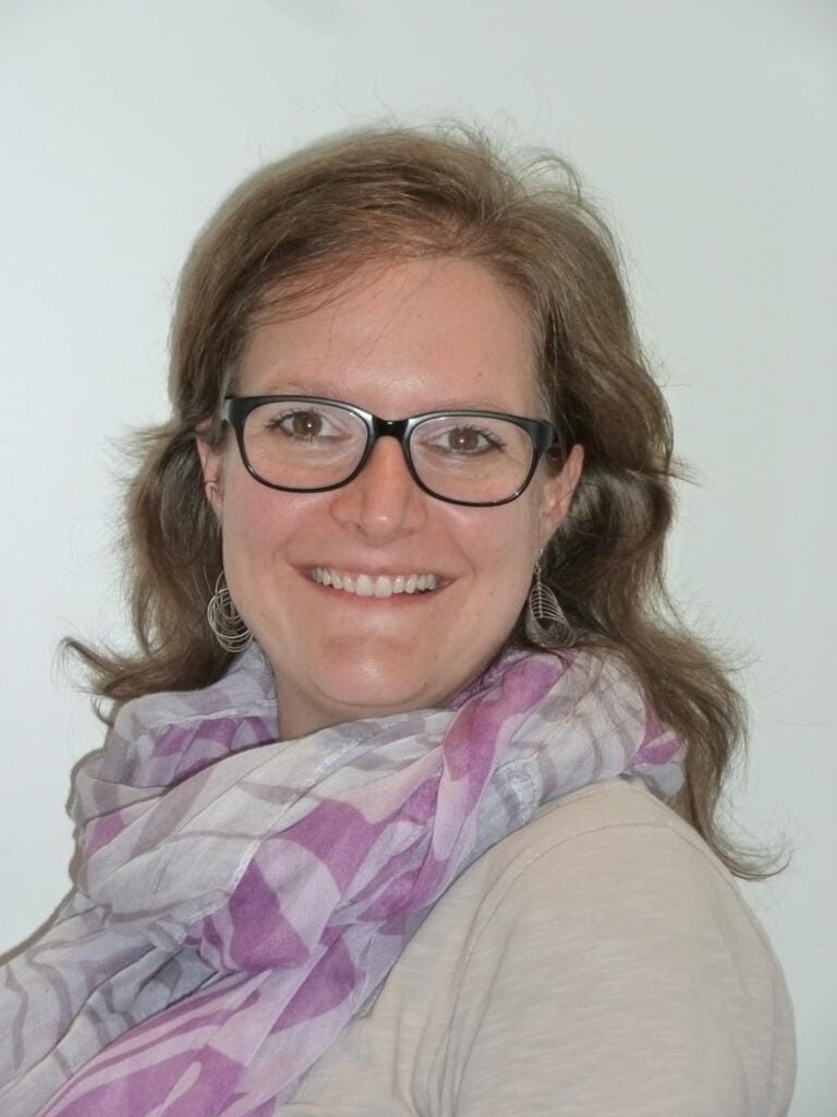 Carmen Burkhalter, eine Frau mit hellbraunem Haar, Brille, beigem Oberteil und hellviolett gemustertem Schal, lächelt vor einem schlichten hellen Hintergrund in die Kamera.