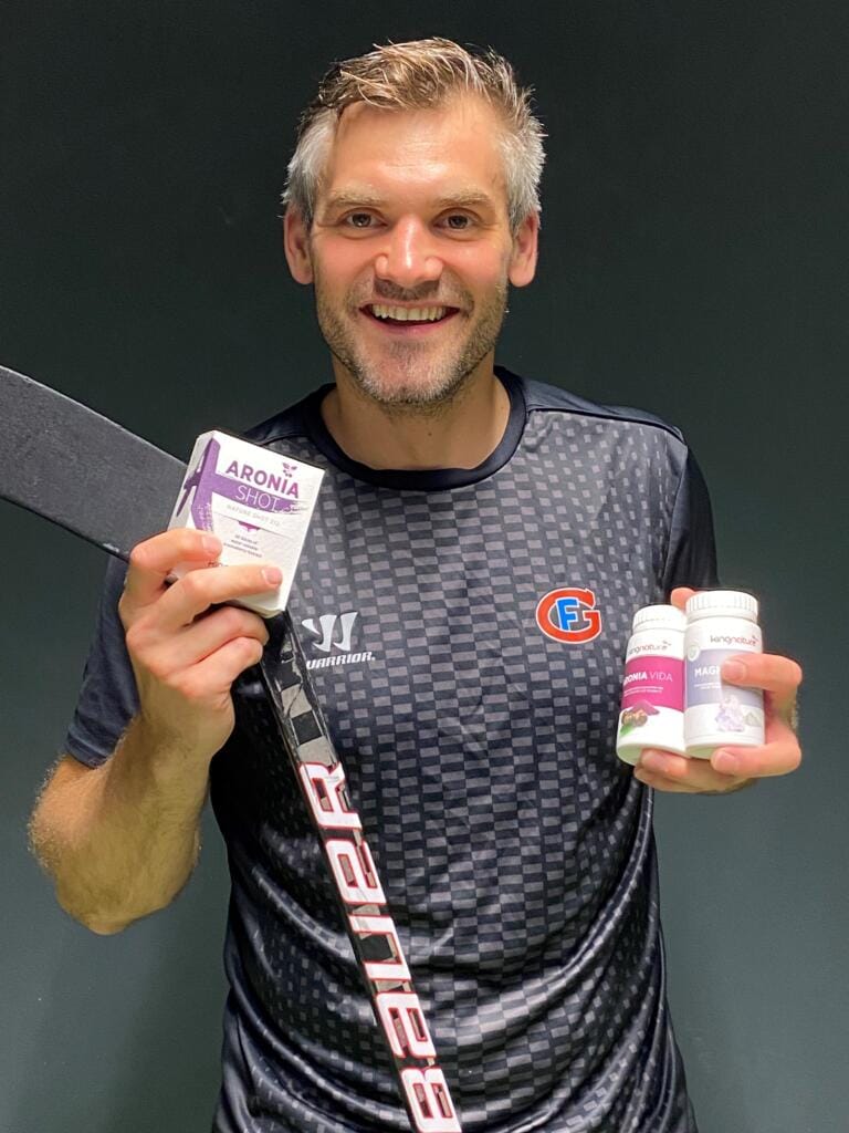 Ein lächelnder Mann in einem schwarzen Sportshirt hält einen Hockeyschläger, eine kleine Schachtel mit der Aufschrift „Aronia Shot“ und zwei Flaschen mit Nahrungsergänzungsmitteln vor einem dunklen Hintergrund und betont damit Gesundheit und Wohlbefinden.
