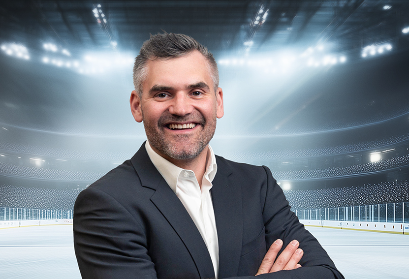 Philippe Furrer, ein lächelnder Mann im Anzug, steht mit verschränkten Armen auf einer leeren, hell erleuchteten Eishockeybahn, im Hintergrund sind Stadionlichter und Sitzplätze zu sehen.