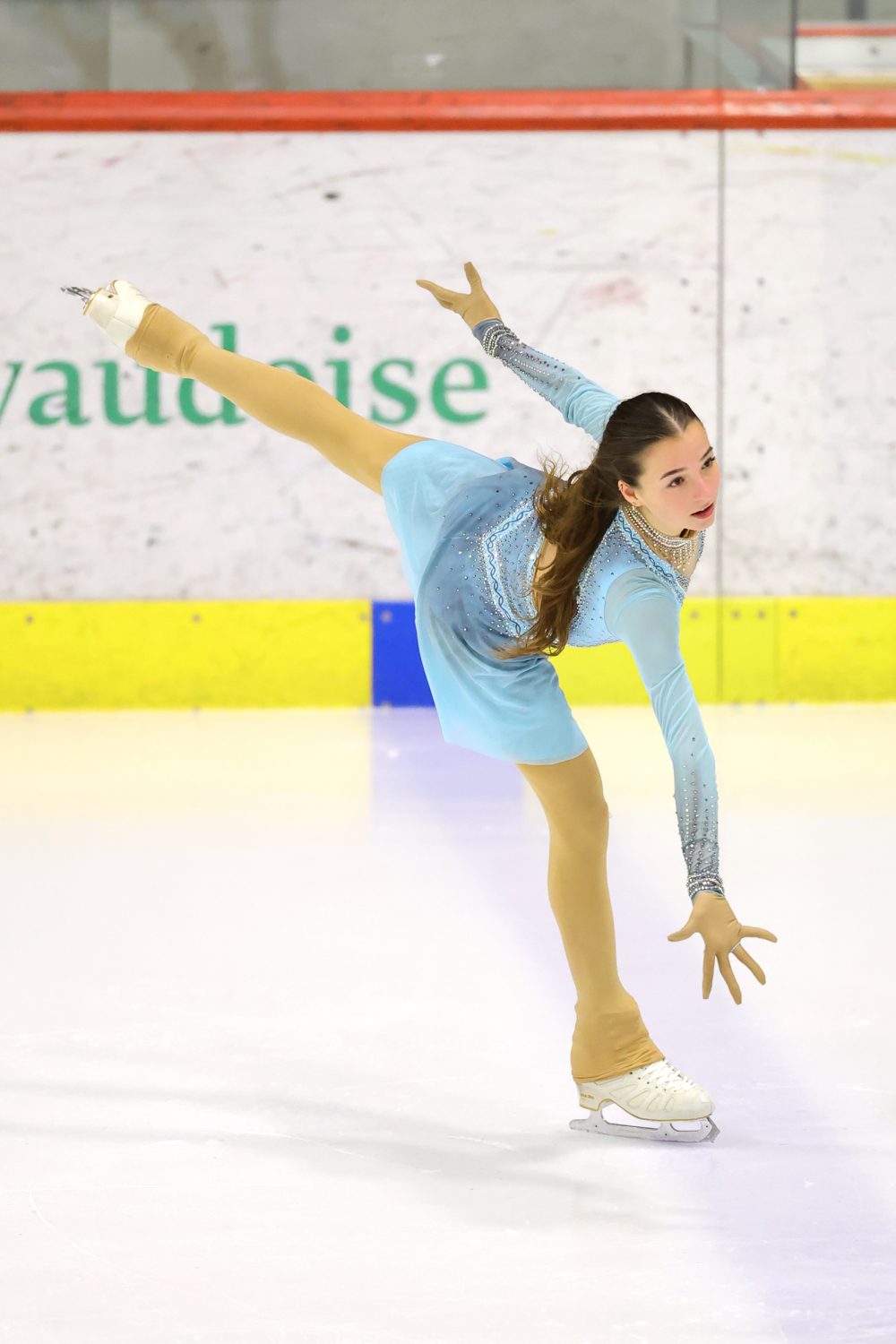 Giada Angelina Molinari, eine junge Eiskunstläuferin in einem blauen Kleid, balanciert auf einem Bein auf dem Eis, wobei sie das andere Bein hoch hinter sich ausstreckt und einen Arm nach vorne streckt.