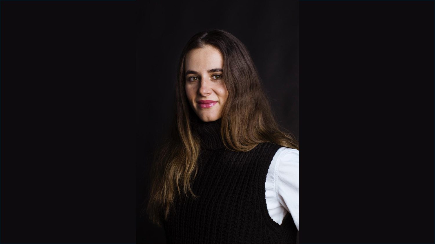 Eine Frau mit langen braunen Haaren trägt eine schwarze Strickweste über einem weißen Hemd und posiert mit einem subtilen Lächeln vor einem dunklen Hintergrund - ein ideales Bild für die Werbung für Eisen Vida.