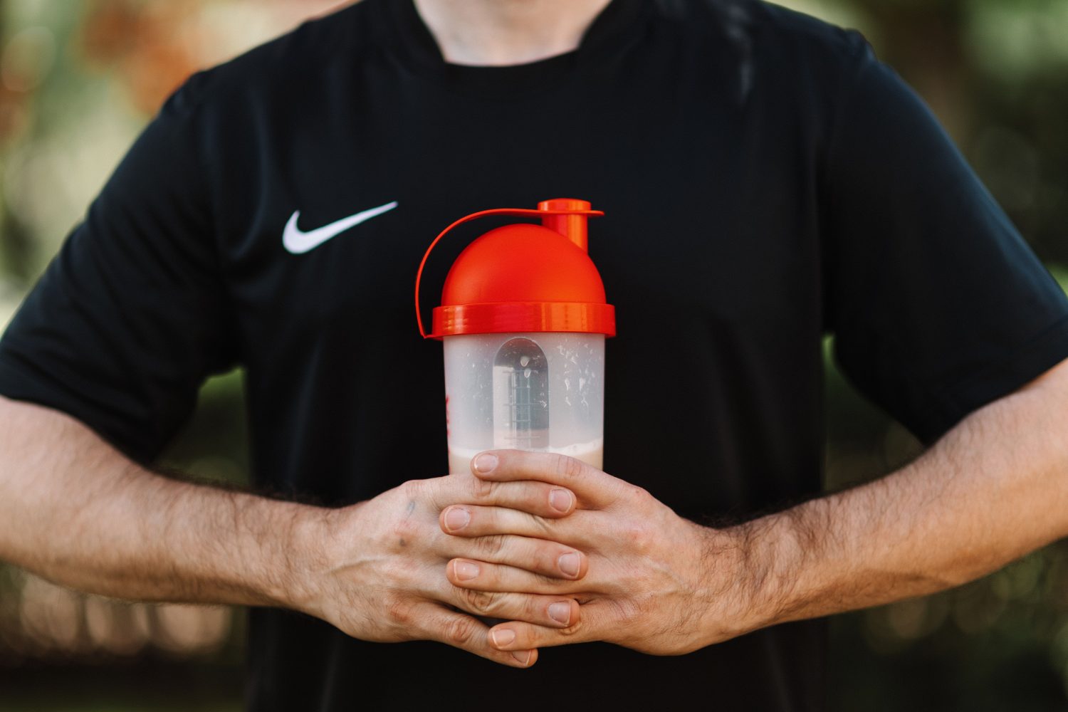 Eine Person in einem schwarzen Nike-Shirt hält mit beiden Händen auf Brusthöhe eine durchsichtige Shaker-Flasche mit rotem Deckel in der Hand. Der Hintergrund ist unscharf, was auf eine Außenaufnahme hindeutet – vielleicht als Teil eines aktiven Lebensstils, um Muskelabbau im Alter und Sarkopenie vorzubeugen.