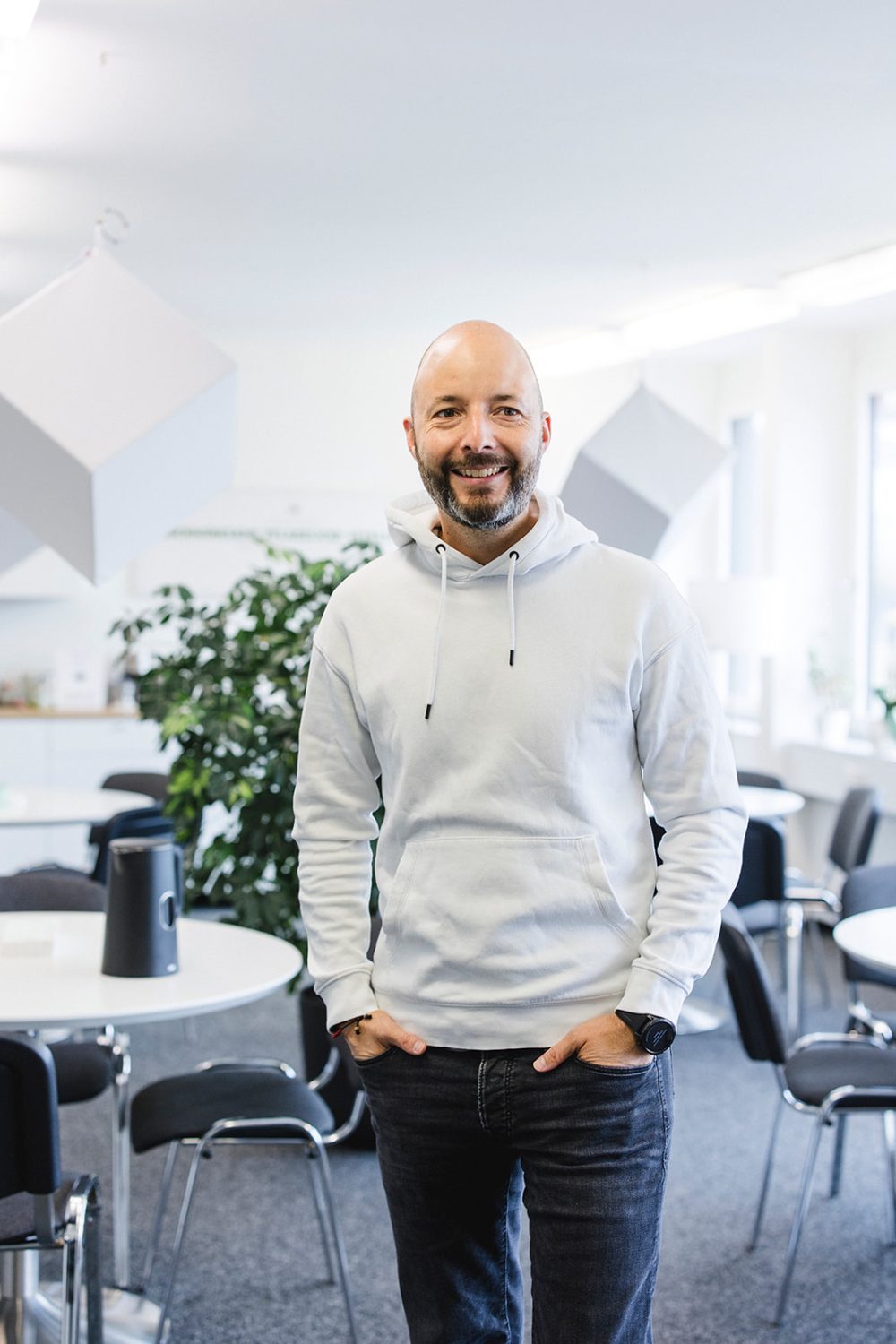 Ein lächelnder Mann mit Bart, einem weißen Kapuzenpullover und Jeans, steht mit den Händen in den Taschen in einem modernen, hellen Büroraum von kingnature mit runden Tischen, Stühlen und Pflanzen im Hintergrund – perfekt für eine Verwaltungsratssitzung.
