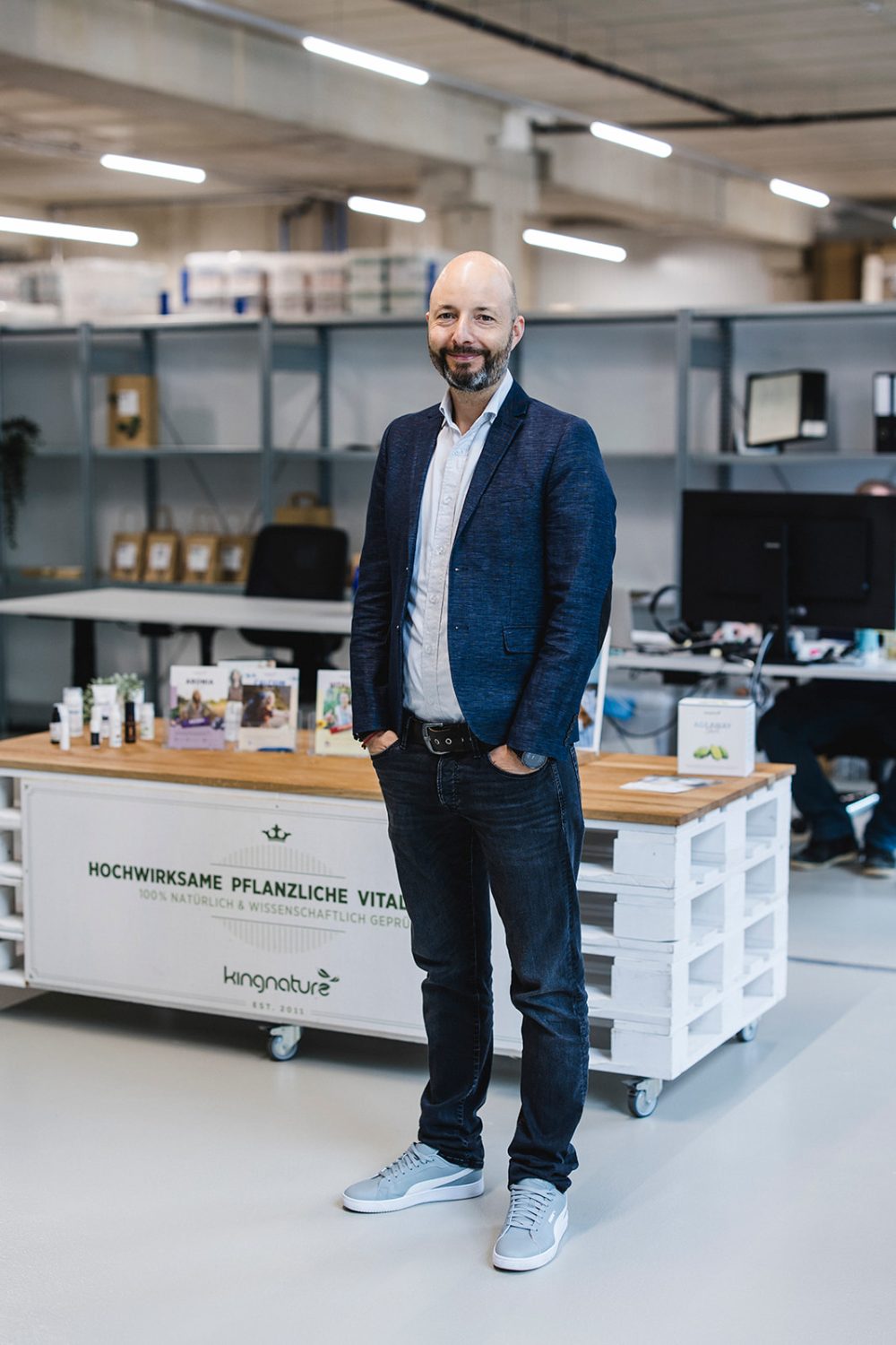 Ein Mann mit Bart, der einen blauen Blazer, ein weißes Hemd, Jeans und hellgraue Turnschuhe trägt, steht lächelnd in einem modernen Büroraum vor einem Ausstellungstisch mit Gesundheitsprodukten von kingnature als Teil des Helden-Verwaltungsrats.