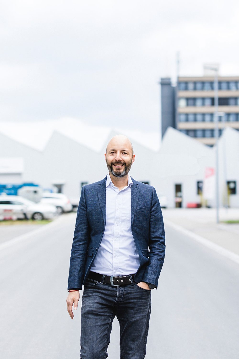 Ein glatzköpfiger Mann mit Bart, einem blauen Blazer und einem weißen Hemd steht mit der Hand in der Tasche auf einer Straße. Hinter diesem möglichen Mitglied des kingnature-Verwaltungsrats befinden sich moderne Gebäude und parkende Autos.