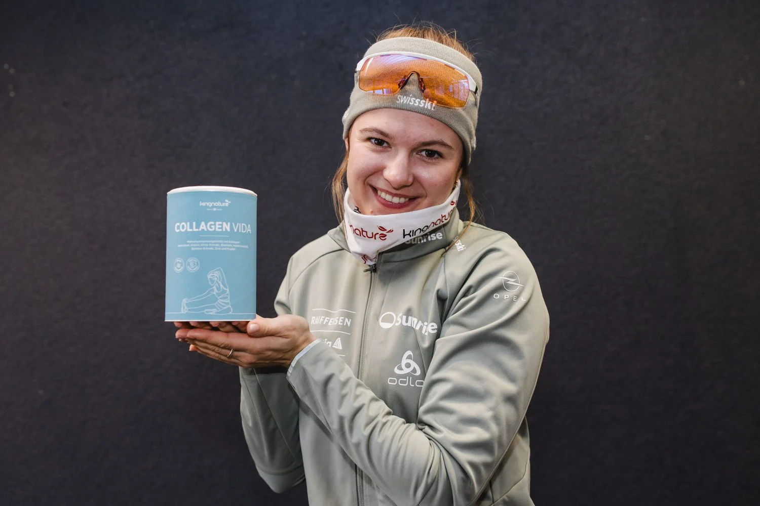 Eine Skifahrerin in einem blau-roten Rennanzug hält einen grünen Ski in der einen Hand und jongliert vor dem dunklen Hintergrund lächelnd mit einer Schachtel und einer Flasche Magnesium Vida mit der anderen Hand.