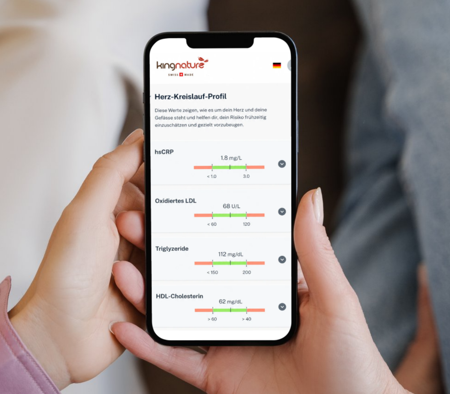 Jemand hält zu Hause ein Smartphone in der Hand, auf dem die Ergebnisse des Herz-Kreislauf-Checks angezeigt werden, darunter farbige Fortschrittsbalken und Werte für hsCRP, oxidiertes LDL, Triglyceride und HDL-Cholesterin.