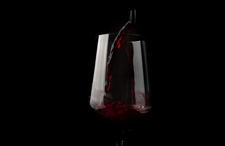Rotwein, der oft als Teil einer ausgewogenen Ernährung genossen wird, wird vor einem tiefschwarzen Hintergrund in ein klares Weinglas gegossen, wodurch die tiefrote Farbe und die dynamische Bewegung der Flüssigkeit besonders gut zur Geltung kommen.
