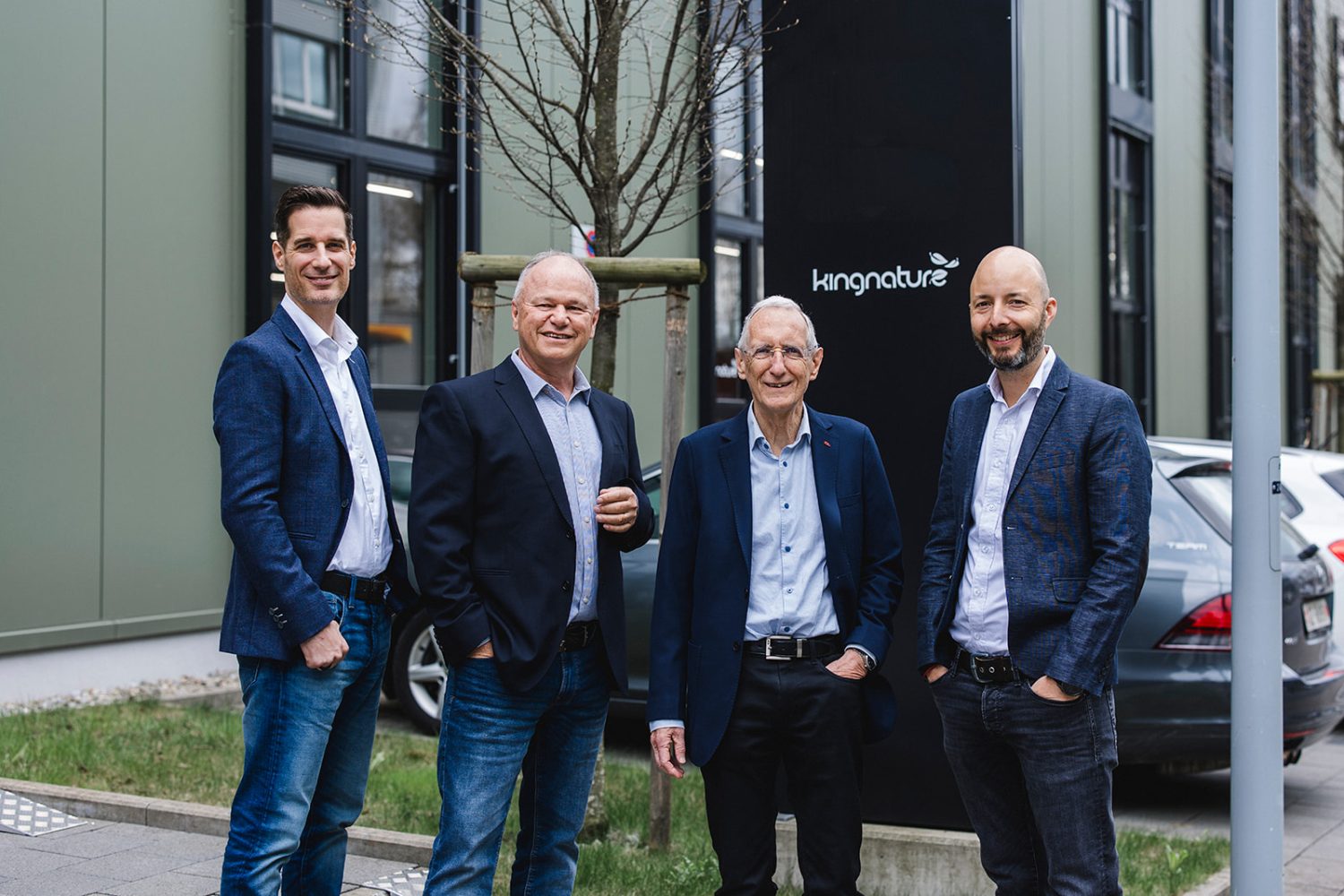 Vier Männer in Business-Casual-Kleidung stehen lächelnd im Freien als Teil des kingnature-Verwaltungsrats und posieren vor einem modernen Gebäude mit großen Fenstern. Im Hintergrund sind Bäume und geparkte Autos zu sehen.