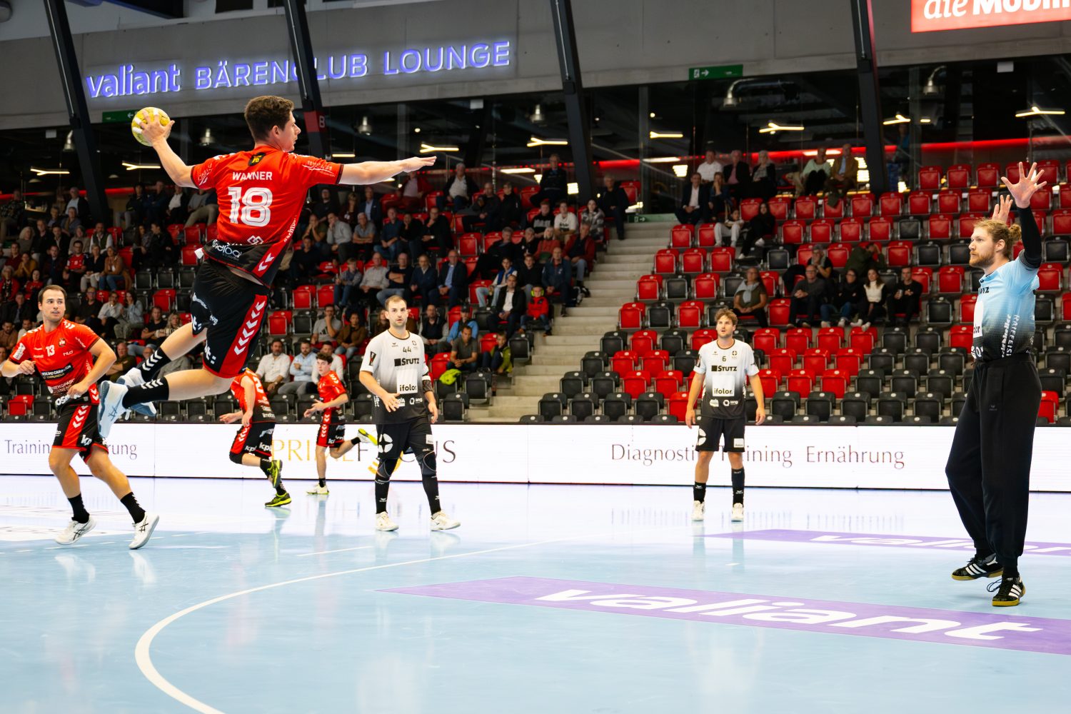 A handball player in a red jersey leaps to shoot the ball while aiming at the goal, as the opposing teams goalkeeper and other players watch attentively in a brightly lit arena with spectators.