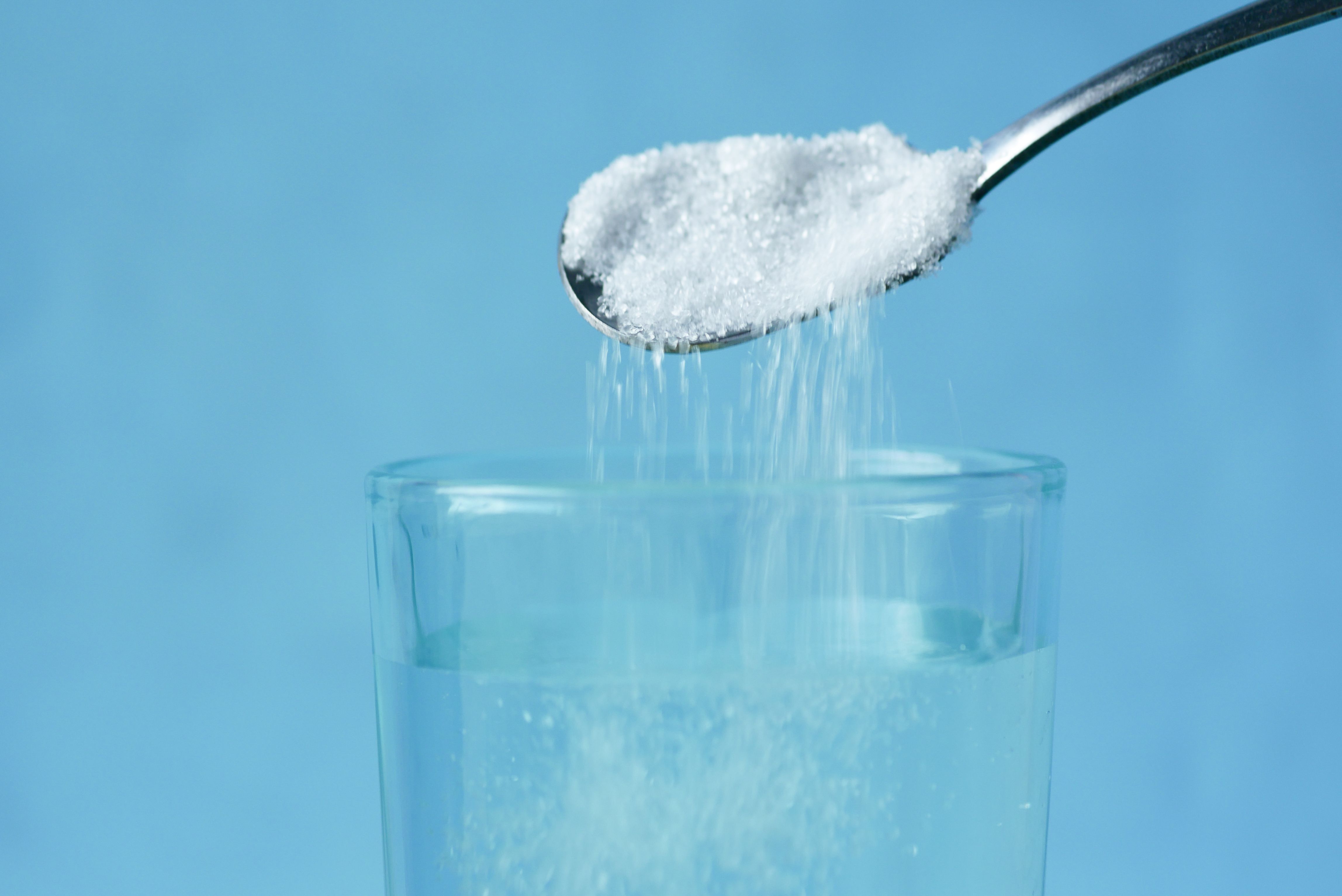 Vor einem blauen Hintergrund wird ein Löffel weißer Kristallzucker in ein klares Glas Wasser geschüttet, um zu verdeutlichen, dass selbst einfache Getränke durch Elektrolyte wie Natrium und Kalium für eine bessere Flüssigkeitszufuhr angereichert werden können.