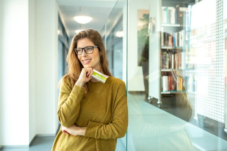 Eine lächelnde Frau mit Brille und senffarbenem Pullover steht in einem modernen Flur und hält eine grüne Plastikkarte an ihr Kinn. Im Hintergrund sind Bücherregale und Glaspaneele zu sehen, die Möglichkeiten zur Konzentrationssteigerung wie mit NADH Vida suggerieren.