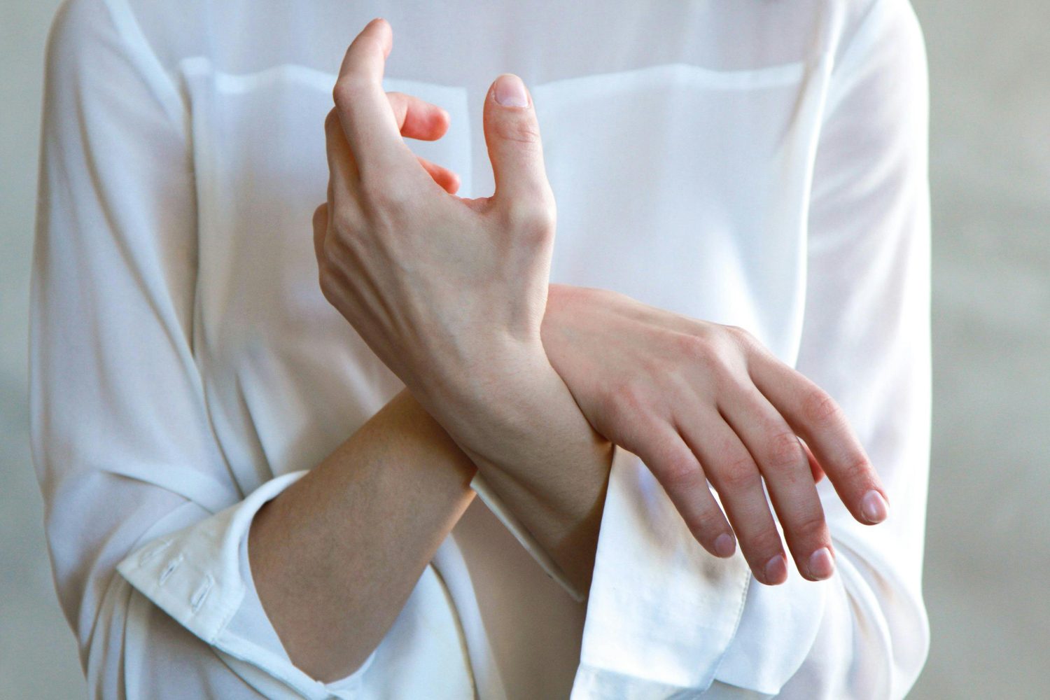 Eine Person in einem weißen, langärmeligen Hemd hält mit der rechten Hand sanft ihr linkes Handgelenk, wobei sich die Finger leicht berühren - was die subtile Besorgnis widerspiegelt, die mit den Symptomen eines B12-Mangels einhergehen kann - vor einem hellen, verschwommenen Hintergrund.