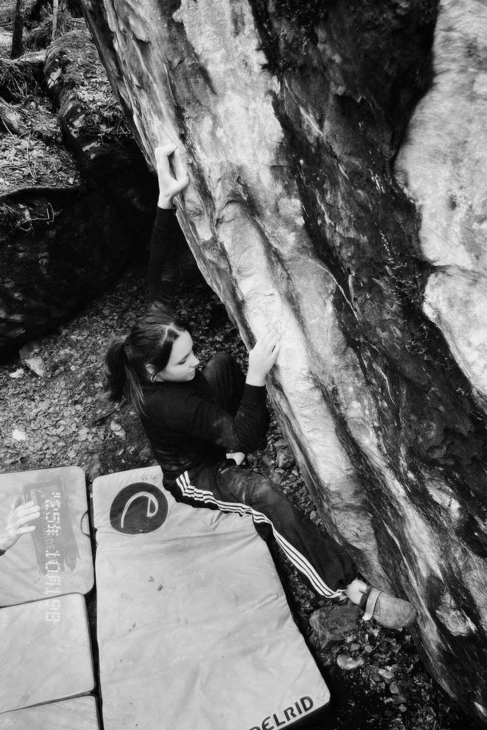 Valentina von Gunten klettert im Freien eine steile Felswand hinauf, wobei sie sich mit einer Hand am Felsen festhält, während sich ihre Füße und die andere Hand festhalten. Zur Sicherheit sind auf dem Boden unter ihr Sturzpads angebracht. Die Szene ist in schwarz-weiß aufgenommen.