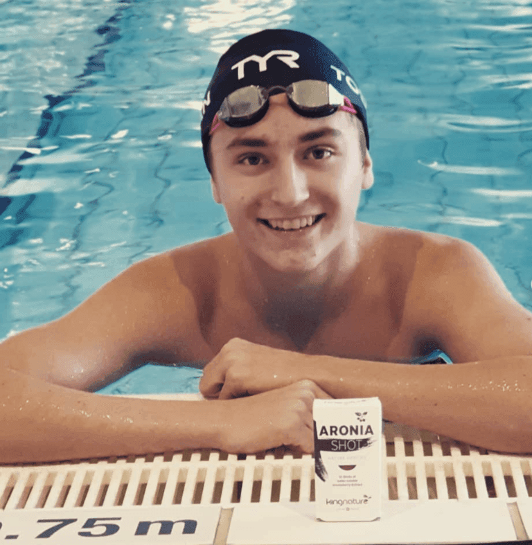 Der Schweizer Schwimmer Marius Toscan lehnt lächelnd mit Schwimmbrille und Badekappe am Rand eines klaren, blauen Pools und hält eine kleine Packung Kingnature Aronia Shot neben sich. Eine Distanzmarkierung zeigt 1,75 m an.