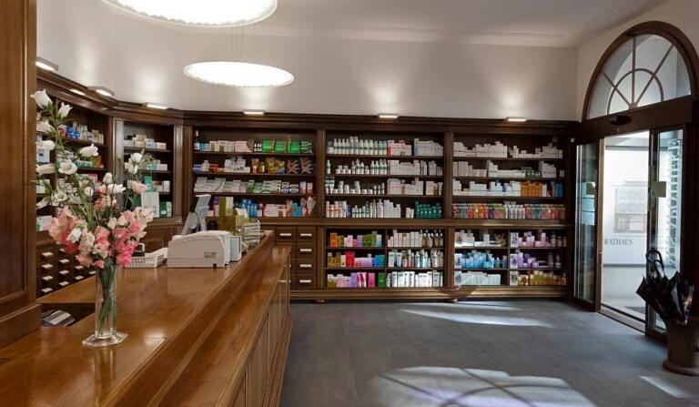 Der saubere, gut beleuchtete Innenraum der Rathaus Apotheke besticht durch Holzregale voller Produkte. Im Vordergrund steht eine Holztheke mit Kasse und einer Vase mit rosa und weißen Blumen, während große Fenster für Tageslicht sorgen.