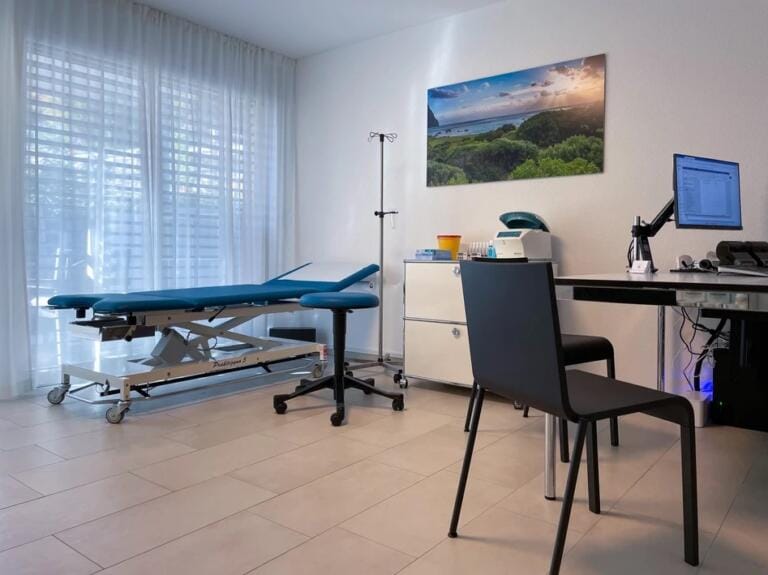 Ein moderner, auf Wellness ausgerichteter Untersuchungsraum mit blauem Untersuchungstisch, Computer auf dem Schreibtisch, schwarzem Stuhl, medizinischer Ausstattung und einem großen Fenster mit transparenten Vorhängen. Ein Landschaftsfoto trägt zur Cordial Health-Atmosphäre bei.