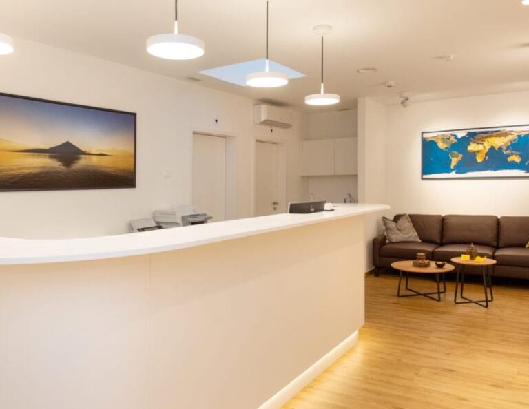 Moderner Empfangsbereich mit geschwungenem weißen Schreibtisch, braunem Sofa, kleinen Tischen und Wandkunst, darunter eine Weltkarte und ein Bergfoto. Warme Beleuchtung und Holzboden schaffen eine einladende Atmosphäre für die Kunden des Swiss Medical AG Health Coaching.