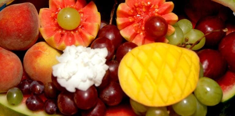 Ein farbenfrohes Arrangement aus verschiedenen Früchten, darunter geschnitzte Wassermelonen, Weintrauben, Pfirsiche, eine gelbe Mango mit einem kreuzförmigen Muster und ein Stück cremeweiße Früchte – perfekt für eine ganzheitliche Ernährungsberatung.