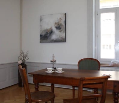 Ein schlichtes Esszimmer mit einem Holztisch, zwei Stühlen, einem Fenster, einer Vase mit Zweigen, zwei Teetassen und einem modernen abstrakten Gemälde an einer hellen Wand schafft einen einladenden Raum für jede Naturheilpraktikerin.