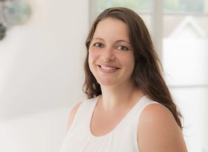 Eine Frau mit langem braunem Haar und einem ärmellosen weißen Oberteil lächelt in die Kamera in einem hellen, sanft beleuchteten Raum mit großen Fenstern und spiegelt die positive Energie wider, die oft in Kinesiologie-AP-Sitzungen zu finden ist.