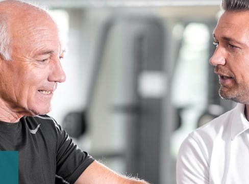 Ein älterer Mann in einem schwarzen Hemd lächelt und unterhält sich mit einem jüngeren Mann in einem weißen Hemd in einem Fitnessstudio oder Fitnesscenter der Vitalwerk AG.