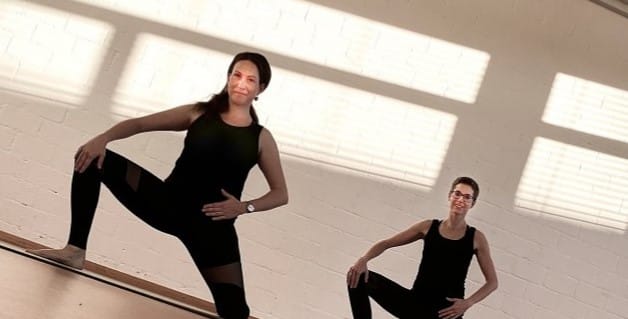 Zwei Personen in schwarzer Trainingskleidung üben eine Pilates-Huttwil-Pose in einem hellen Raum mit weißen Backsteinwänden und großen Fenstern, die Schatten werfen. Beide stehen mit gebeugten Knien und den Händen in den Hüften da und lächeln.