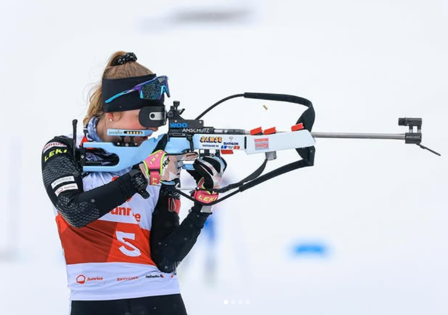 Eine Biathletin in Wintersportausrüstung zielt während eines Wettkampfs mit einem Gewehr. Sie trägt eine rote Startnummer mit der Nummer 5, schwarze Handschuhe, eine Sonnenbrille und ein Stirnband vor einem verschneiten Hintergrund.