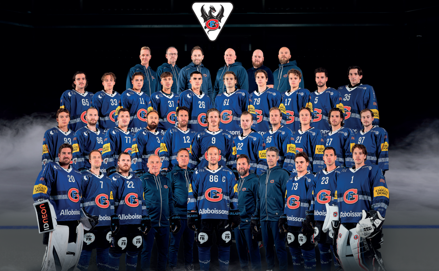 Eine große Gruppe von Schweizer Eishockeyspielern und -mitarbeitern in blauen Uniformen posiert gemeinsam für ein Mannschaftsfoto vor einem dunklen Hintergrund, über dem das Teamlogo des HC Fribourg-Gottéron zu sehen ist.