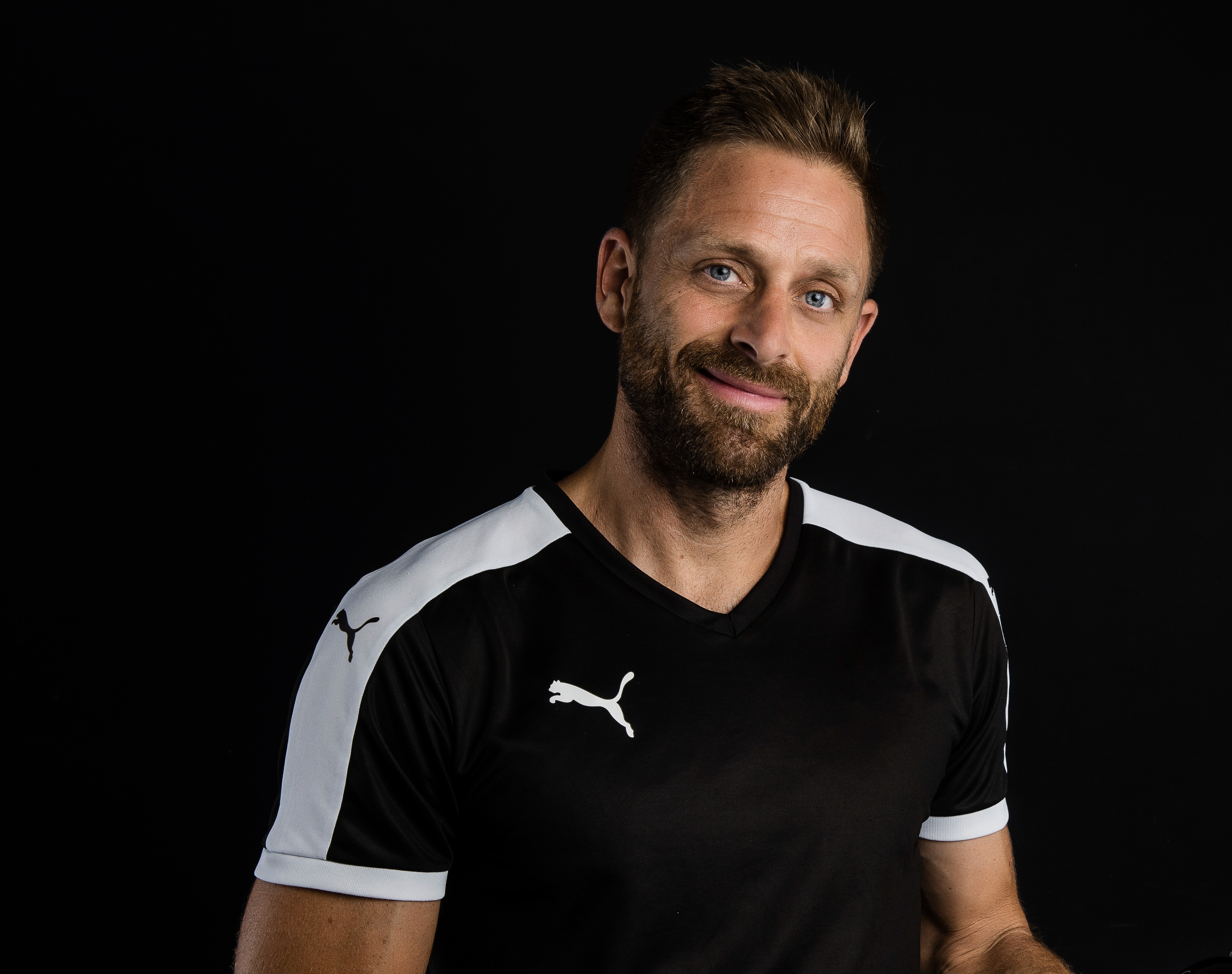 Ein Mann mit kurzen braunen Haaren und Bart lächelt, trägt ein schwarz-weißes Puma-Sportshirt, steht vor einem schlichten schwarzen Hintergrund und hält eine Flasche Amino Vida.