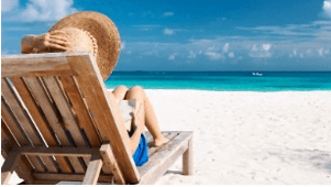 Eine Person entspannt sich auf einem hölzernen Liegestuhl an einem Sandstrand, trägt einen großen Sonnenhut und trägt Sonnencreme auf, während sie unter einem strahlend blauen Himmel mit vereinzelten Wolken auf das türkisfarbene Meer blickt.