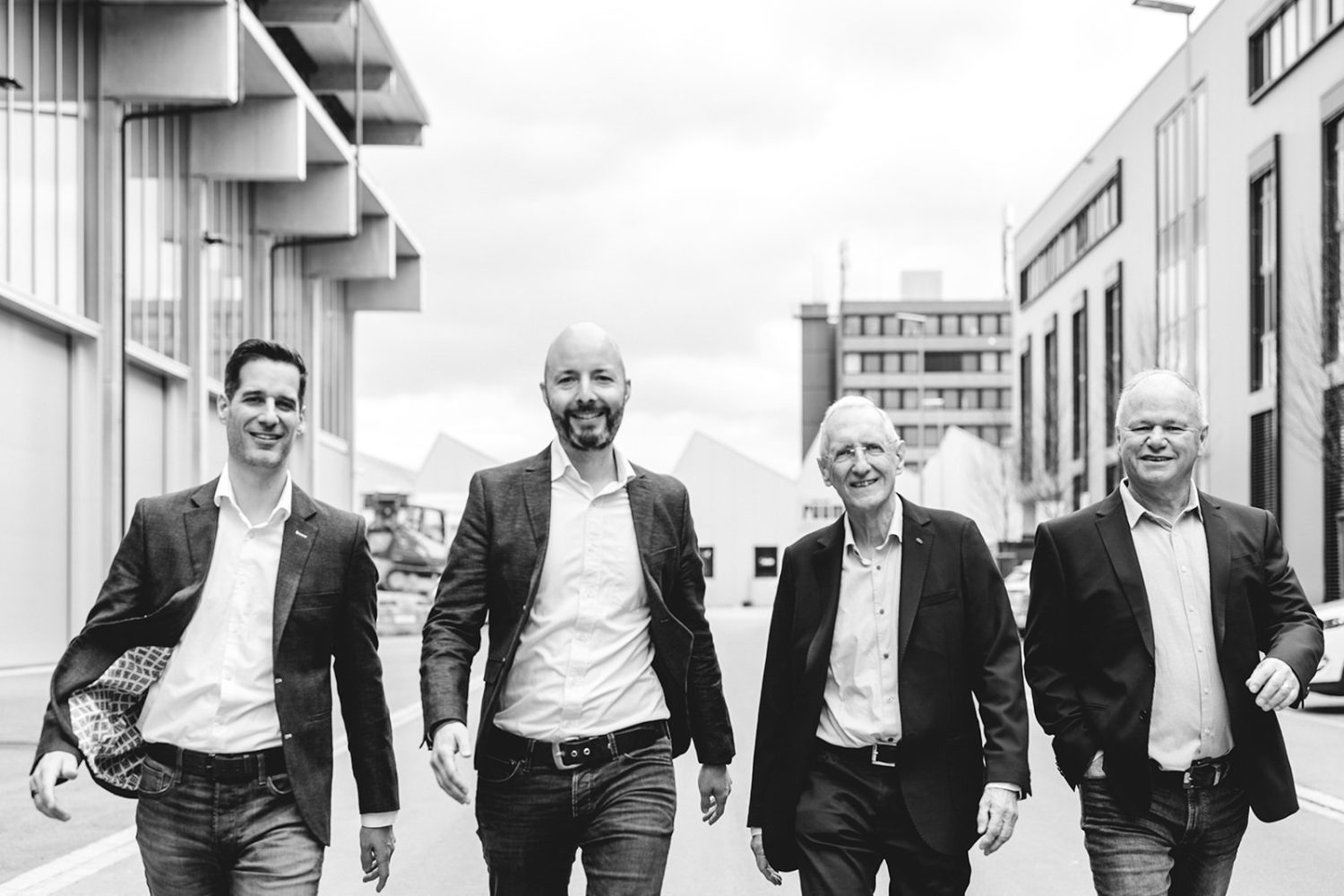 Vier Männer in Businesskleidung, darunter Mitglieder des kingnature-Verwaltungsrats, gehen lächelnd und selbstbewusst zwischen modernen Gebäuden umher. Das Schwarz-Weiß-Foto fängt eine professionelle und freundliche Atmosphäre ein.