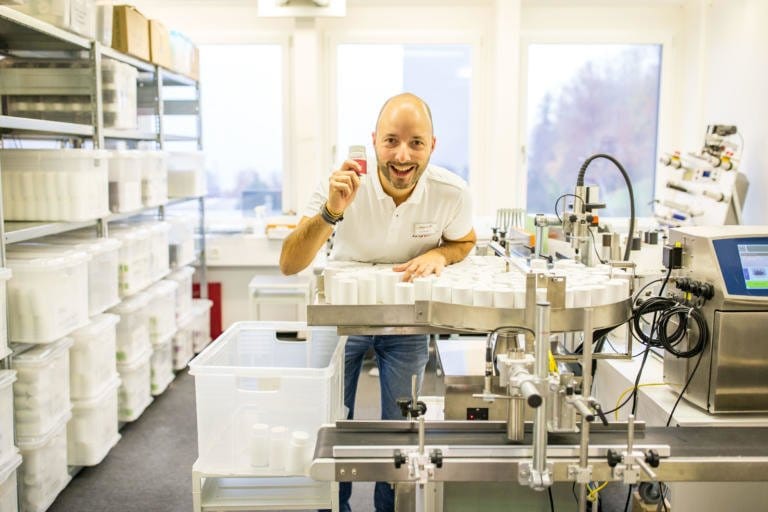 Ein lächelnder Mann mit einem kleinen Behälter in der Hand steht in einem hellen Labor voller Geräte und Lagerbehälter, was an eine Produktions- oder Verpackungsumgebung bei kingnatures Tag der offenen Tür erinnert.