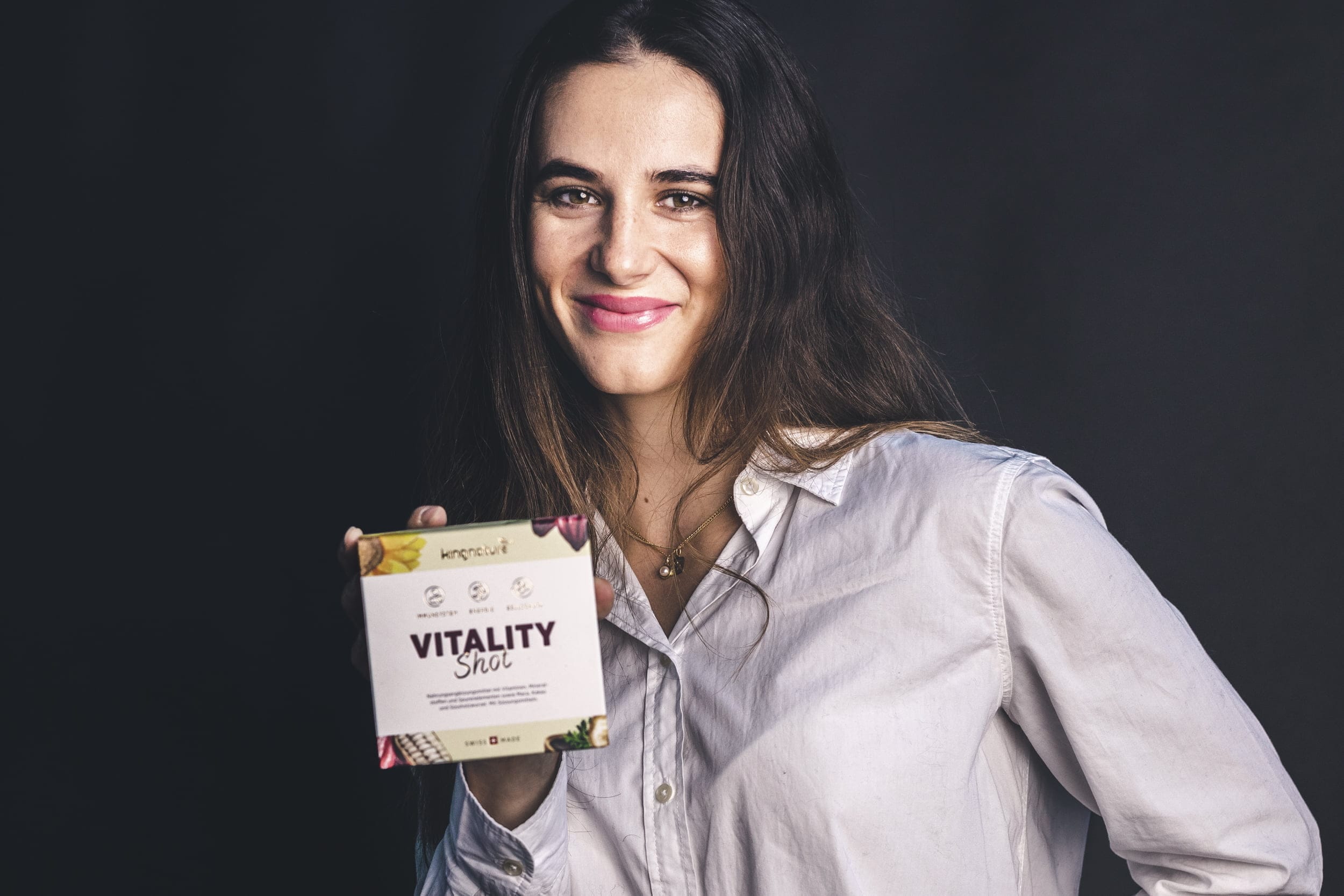 Talina Gantenbein, mit langen braunen Haaren und einem weißen Hemd mit Knöpfen, lächelt, während sie vor einem dunklen Hintergrund eine Schachtel mit der Aufschrift „Vitality Shot“ hält.