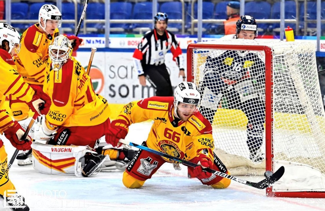 A hockey player in a yellow and red uniform dives in front of the goal, attempting to block the puck, while the goalie and another teammate defend. An opposing player in blue stands near the net as referees observe in the background.