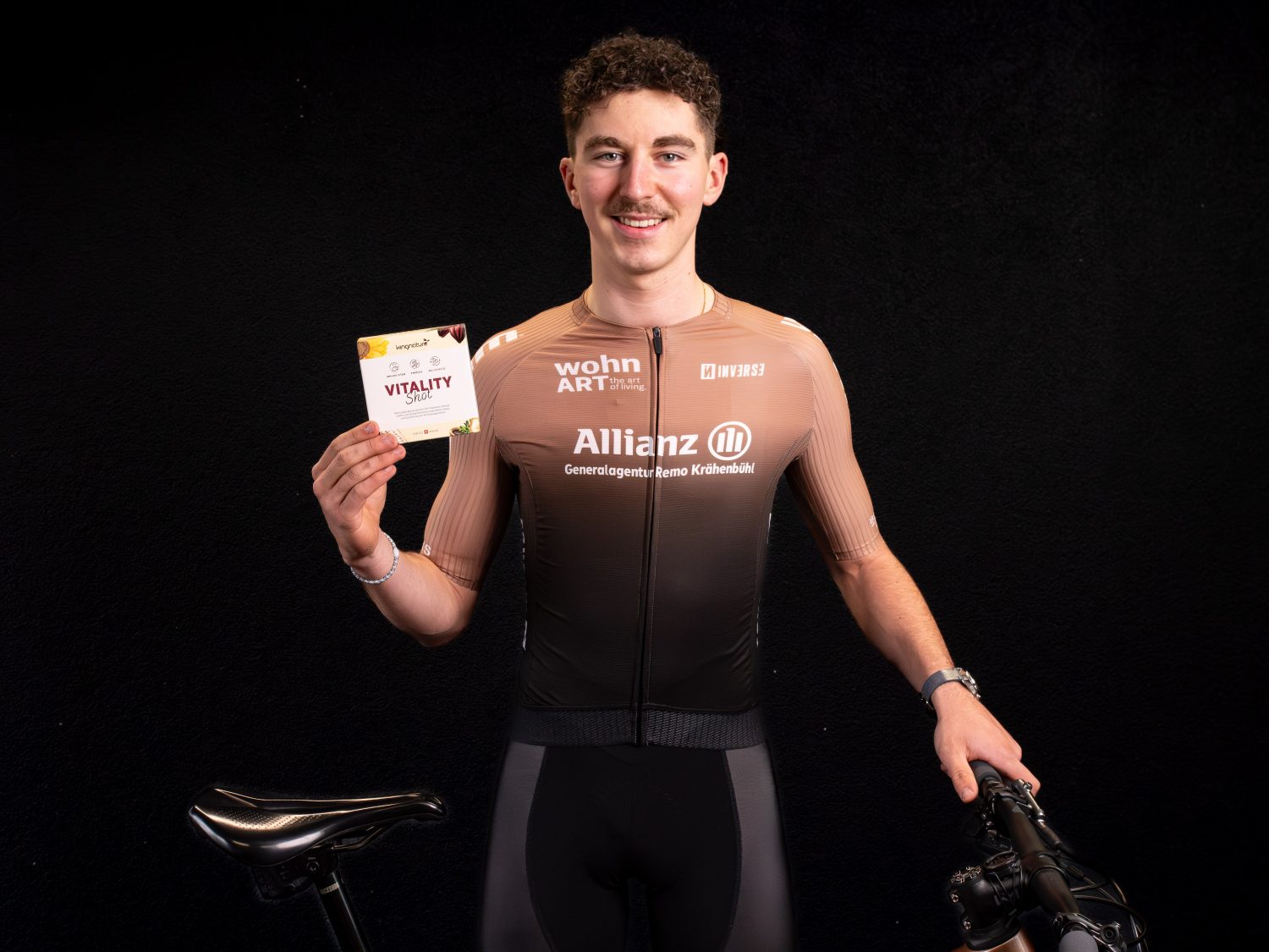Timon Minder, ein männlicher Radfahrer in einem braun-schwarzen Trikot, steht neben einem Fahrrad, lächelt und hält einen Vitality-Ernährungsriegel vor einem dunklen Hintergrund hoch.