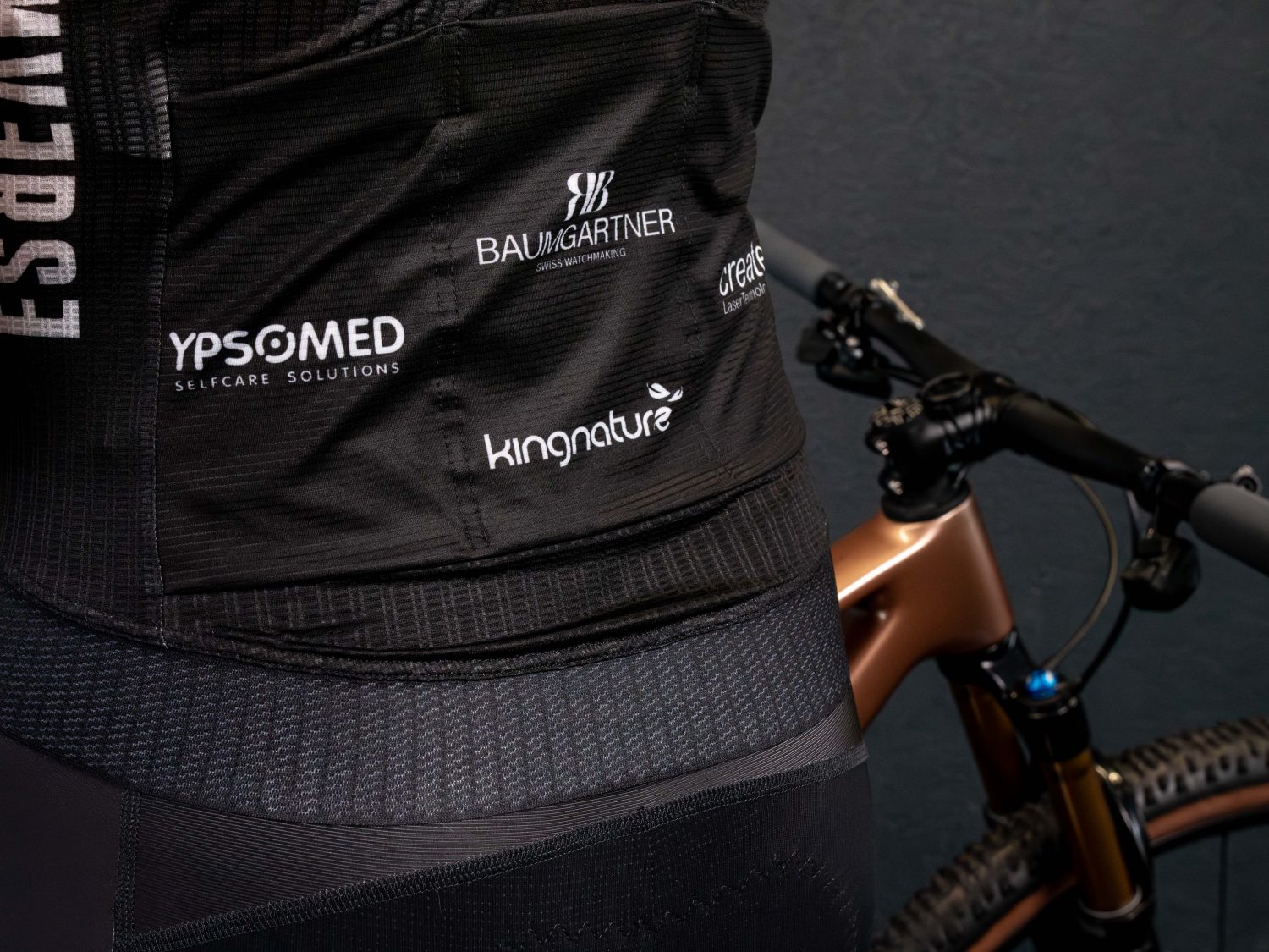 Eine Nahaufnahme von Timon Minders Rücken zeigt Sponsorenlogos auf einem schwarzen Trikot, während im Hintergrund der Lenker und das Vorderrad eines bronzenen Fahrrads zu sehen sind.