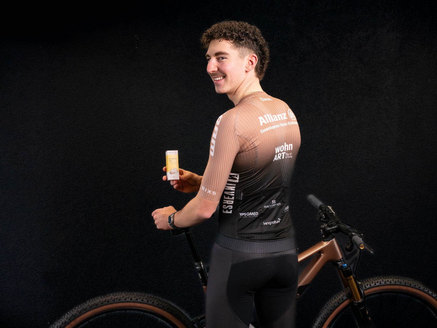 Timon Minder, ein Radfahrer in einem schwarz-braunen Fahrraddress, steht neben einem Fahrrad, lächelt und hält eine kleine gelbe Tube oder Flasche vor einem schwarzen Hintergrund in der Hand.