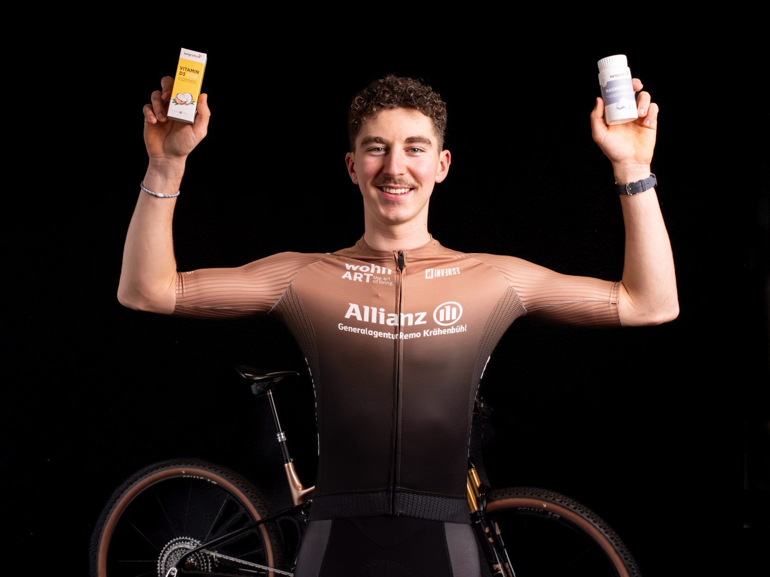 Timon Minder, ein männlicher Radfahrer in einem braunen Radtrikot, steht mit erhobenen Armen, in der einen Hand eine kleine gelbe Schachtel und in der anderen eine weiße Flasche haltend, lächelnd vor einem schwarzen Hintergrund, hinter dem ein Fahrrad teilweise sichtbar ist.