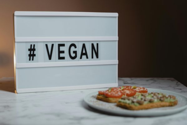 Ein Leuchtkasten mit der Aufschrift #VEGAN steht auf einer Marmorplatte neben einem Teller mit Sandwiches, die mit Tomatenscheiben und Samen belegt sind - der perfekte Treibstoff für alle, die bei der Aufnahme von Kreatin in ihren Speiseplan auf Dosierung und Wirkung achten.